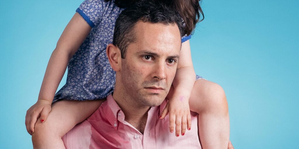 Photo of Adam Lazarus wearing a pink button up with a child sitting on his shoulders in front of a blue background