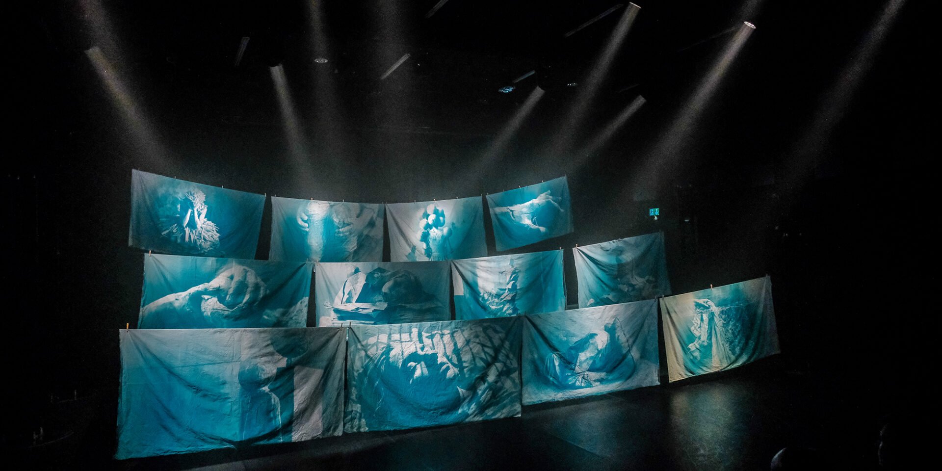 A large stage with piecies of fabric died blue hanging in three rows with lights shining down on them.