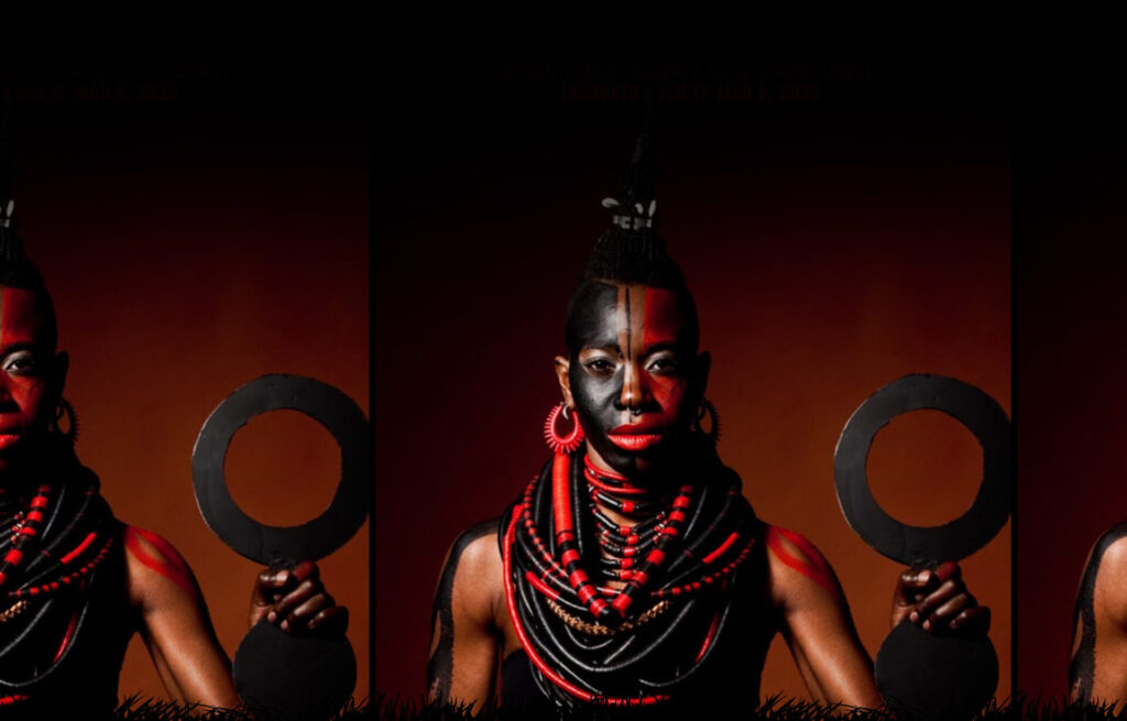 dbi young, in traditional attire, hair tied tall, with face painted in red and black, wearing black and jewel beads reminiscent of an armour, regally stares at the camera.