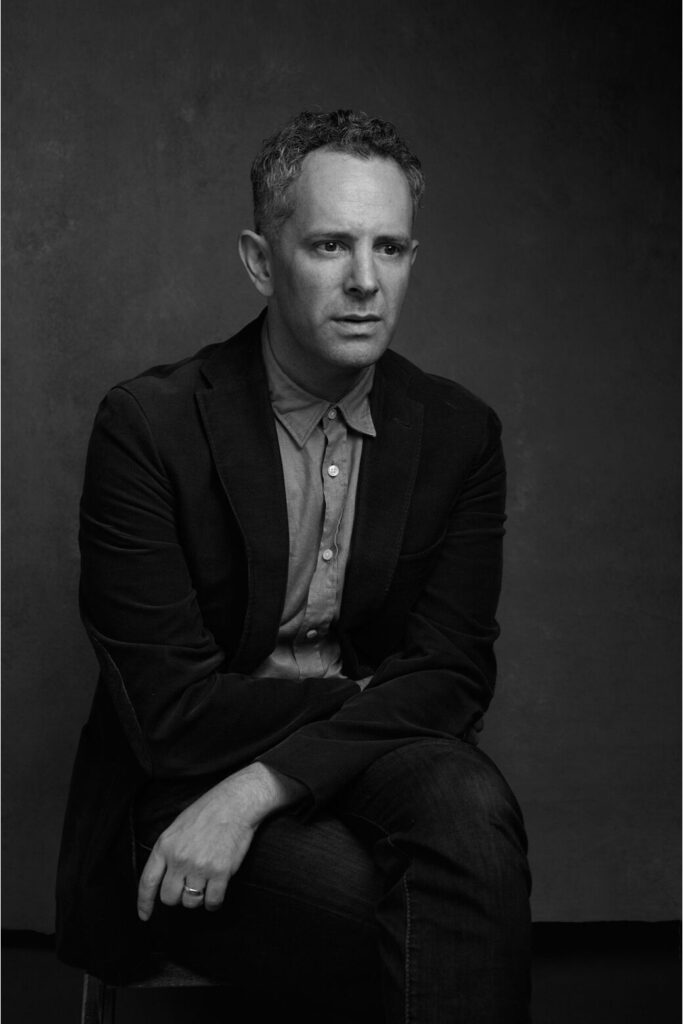 black and white photo of Adam looking stoic and a bit confused sitting with his arms gently crossed in front of him.
