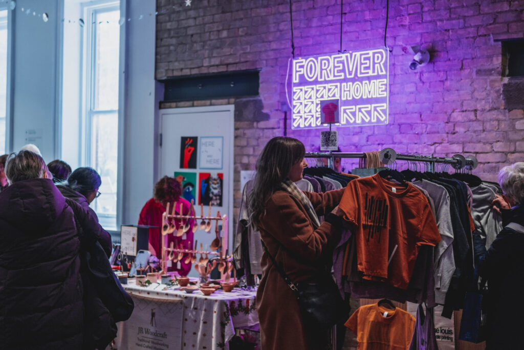 A shopper looking at clothing on a rack with handmade wooden spoons on a table behind them in the gallery space. A glowing purple neon sign reads forever home in front of a brick wall.