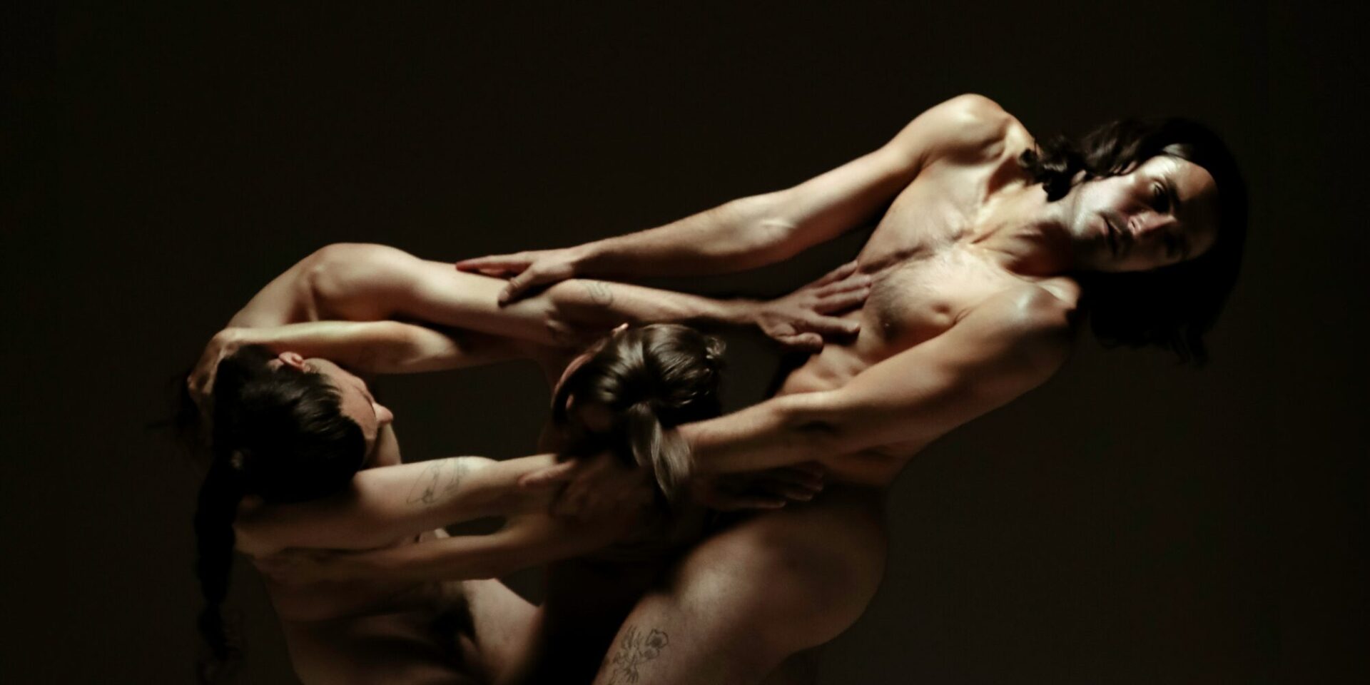 three dancers are mid-movement, fully nude. They are lit in dramatic dark and bright lighting like in a baroque painting. Their arms are woven together and the three people appear like they are one form.