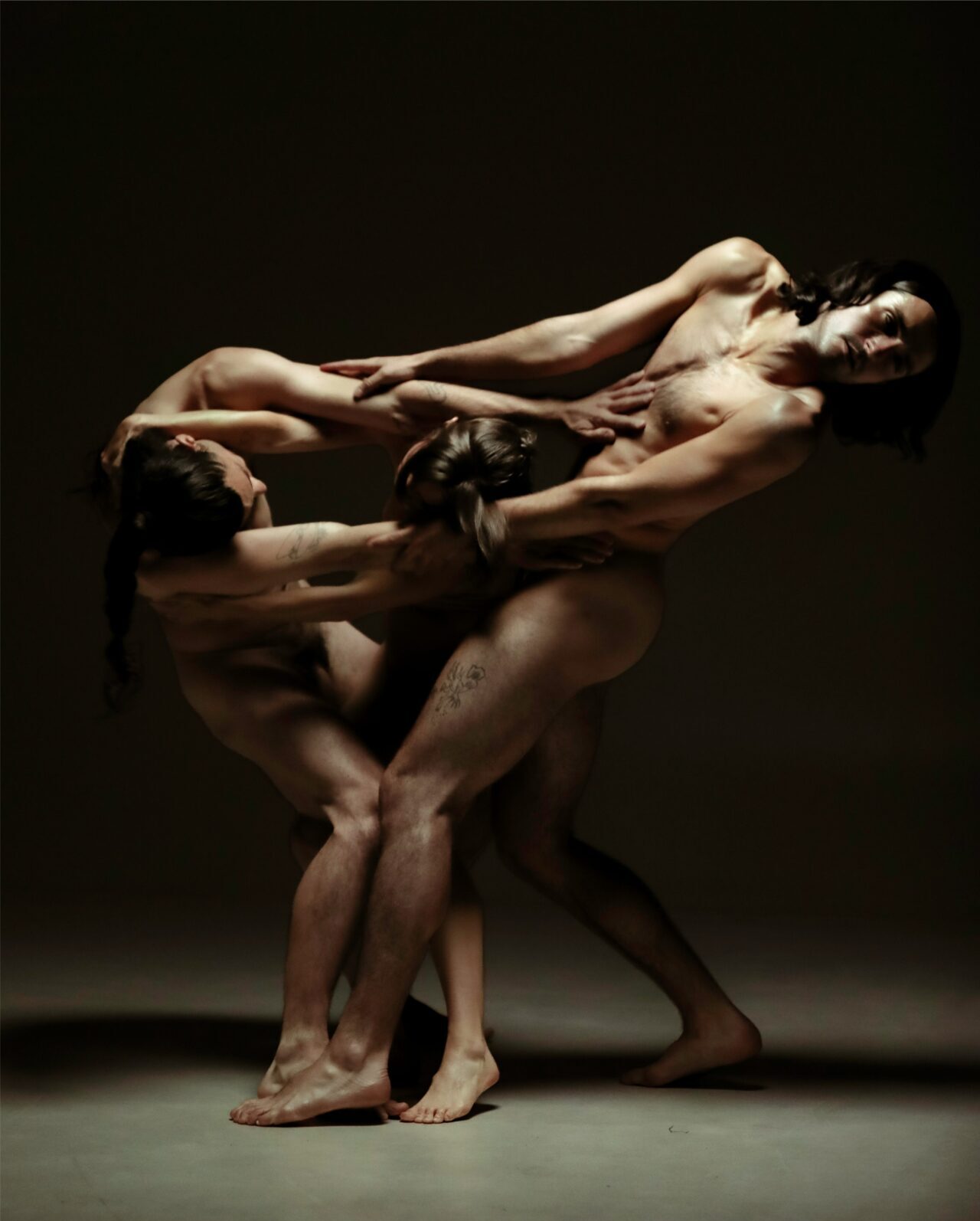 three dancers are mid-movement, fully nude. They are lit in dramatic dark and bright lighting like in a baroque painting. Their arms are woven together and the three people appear like they are one form.